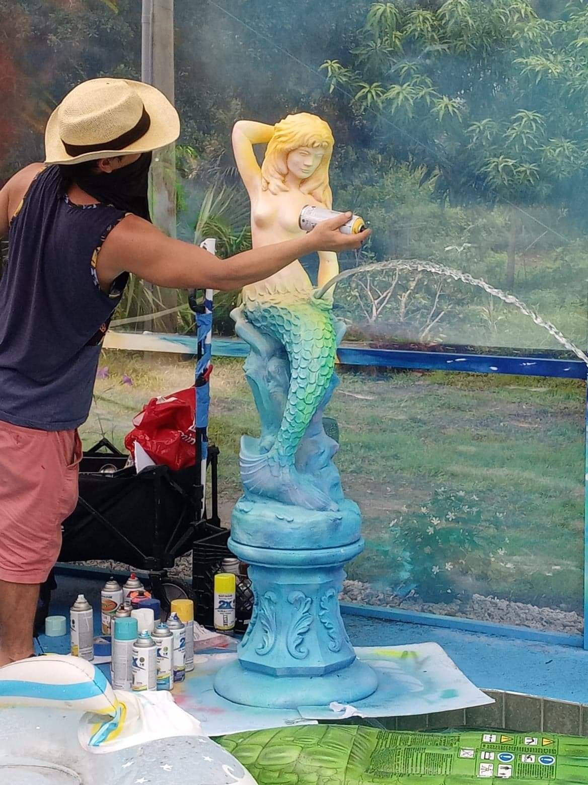 Artist spray painting a mermaid fountain statue in a pool enclosure